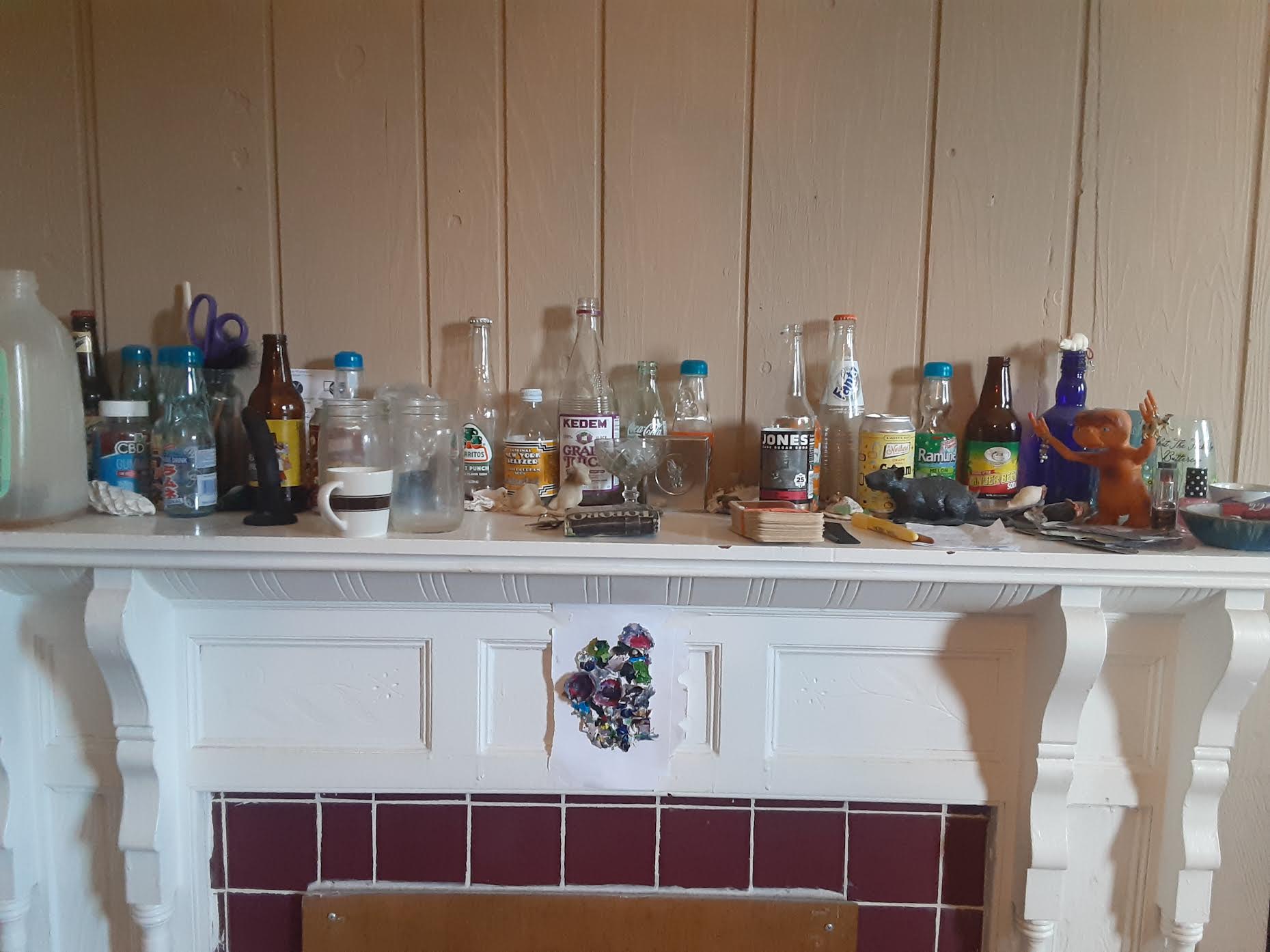 defunct fireplace mantle with arrayed objects on it, including many bottles, a dildo, shells, and hard-to-make-out detritus, can click this image to enter a page that says "come in!